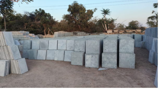 Stacks of grey Kota stone slabs neatly arranged in an open yard with trees and a clear sky in the background.